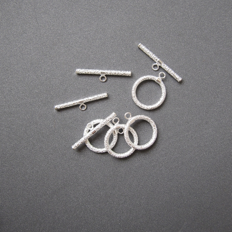 May include: Silver toggle clasps with a textured finish. The clasps are in the shape of a bar and a circle. There are five clasps in total.