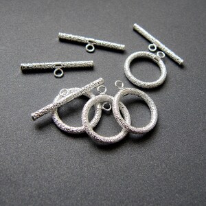 May include: Silver toggle clasps with a textured finish. The clasps are in the shape of a bar and a circle. The bar clasps have a small loop on each end. The circle clasps have a small loop on one side.