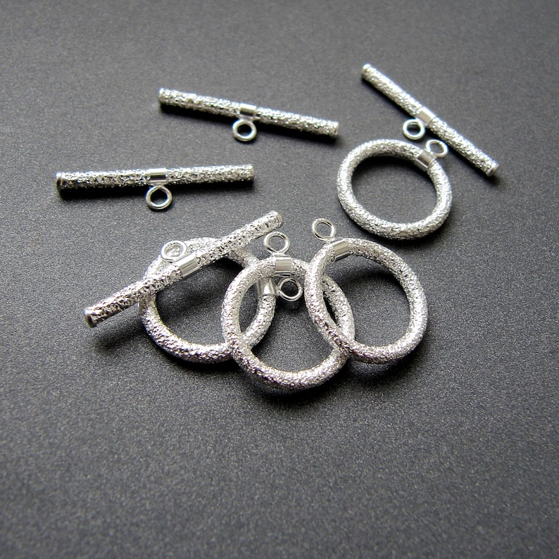May include: Silver toggle clasps with a textured finish. The clasps are in the shape of a bar and a circle. The bar clasps have a small loop on each end. The circle clasps have a small loop on one side.