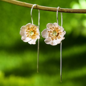 May include: Silver drop earrings featuring flower-shaped pendants. The earrings have white petals with gold-colored accents and a green gemstone center. The earrings are suspended from thin silver wires, creating a delicate and elegant design.