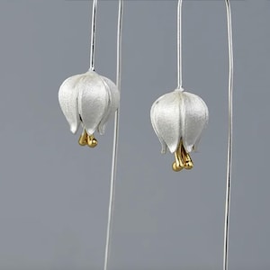 Op de afbeelding: Een paar zilveren oorbellen met witte, bloemvormige bedels en gouden accenten. De oorbellen hangen aan dunne zilveren draden.