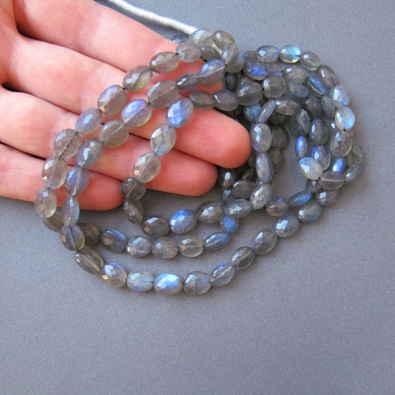 May include: A strand of oval-shaped, faceted labradorite beads in shades of grey with blue iridescent flashes. The beads are strung on a clear thread and are approximately 10mm in size.