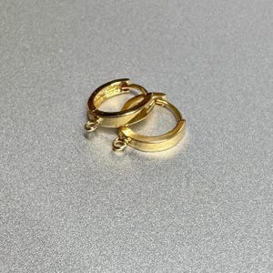 Può includere: Due orecchini a cerchio color oro con un piccolo anello nella parte inferiore per attaccare ciondoli o altri abbellimenti. Gli orecchini hanno un design semplice e classico e una finitura lucida. Gli orecchini sono esposti su una superficie argentata.