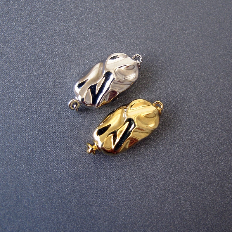 Op de afbeelding: Twee metalen sluitingen voor het maken van sieraden. E&eacute;n is zilver en de andere is goud. Beide hebben een textuur en een organische vorm.