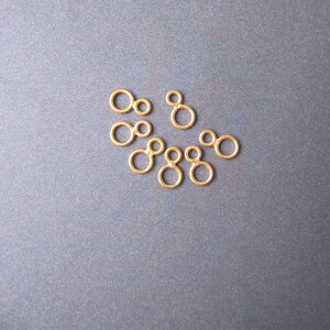 May include: A set of eight gold-colored metal jump rings, each with two connected circles. The rings are arranged in a loose cluster on a gray surface.