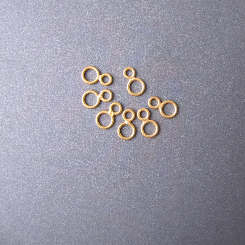 May include: A set of eight gold-colored metal jump rings, each with two connected circles. The rings are arranged in a loose cluster on a gray surface.