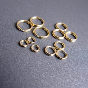 Puede incluir: Una colección de anillas de salto de metal en tono dorado de varios tamaños. Estos fornituras circulares para joyería presentan un diseño dividido, lo que permite abrirlas y cerrarlas para conectar componentes en la creación de joyas.