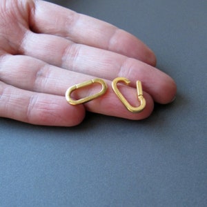 May include: Two gold-colored metal clasps, one oval-shaped and one with a hook, are shown on a hand.