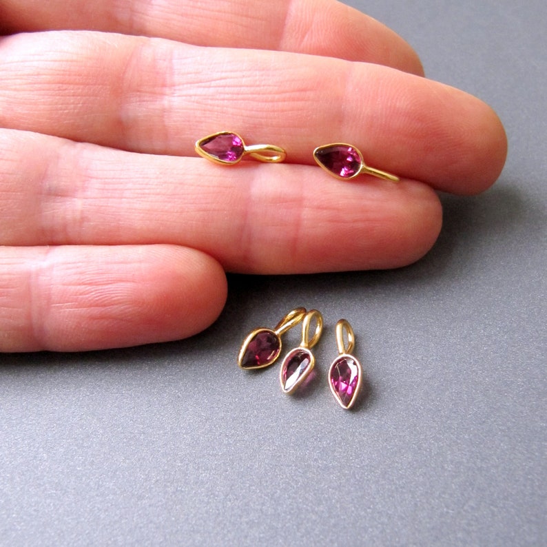 Op de afbeelding: Vijf goudkleurige bedels met roze peervormige edelstenen. De bedels zijn ontworpen om aan kettingen of armbanden te worden toegevoegd.