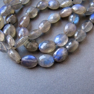 May include: A strand of oval-shaped, faceted labradorite beads with a blue iridescent sheen. The beads are strung on a clear thread and are arranged in a loose, overlapping pattern.