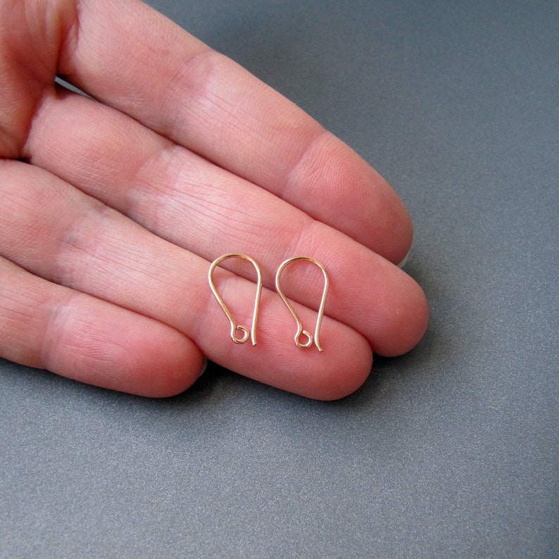 May include: Two gold-toned earring hooks, also known as French hooks, are shown on a hand. The hooks are made of metal and have a simple, elegant design.