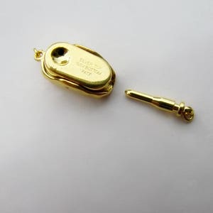 Op de afbeelding: Een gouden metalen hanger met een zilveren bovenkant en een klein gaatje in het midden. De hanger heeft de tekst "SILVER TOP GSV BOTTOM PAT.P" gegraveerd. Een aparte gouden metalen staaf met een afgerond uiteinde is ook afgebeeld.