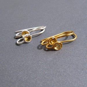 May include: Two rings, one silver and one gold, each featuring two round, yellow gemstones. The silver ring has a delicate, intertwined design, while the gold ring has a similar, yet distinct, style. The rings are displayed on a gray surface.
