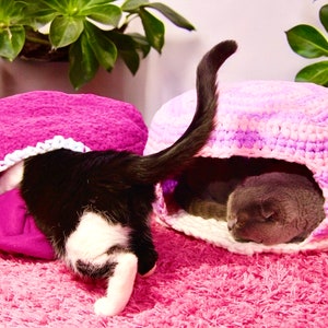 May include: Two cats resting in crocheted cat beds. One cat is black and white and is peeking out of a pink bed. The other cat is gray and is inside a pink and purple striped bed.