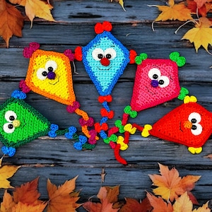 May include: Five colorful crocheted kites with smiling faces, connected together with colorful yarn, on a wooden background with autumn leaves.