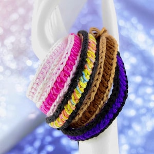 Könnte beinhalten: Ein Stapel bunter gehäkelter Armbänder in Rosa, Weiß, Gelb, Braun, Schwarz und Lila.