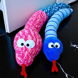 May include: Two handmade crochet snakes. One is pink with white eyes and a red tongue. The other is blue with white eyes and a red tongue. The snakes are lying on a black surface.