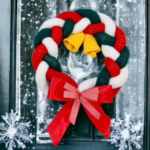 Puede incluir: Una corona de Navidad de ganchillo con hilo rojo, blanco y verde, decorada con un lazo rojo y campanas amarillas.