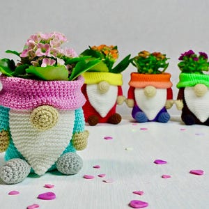 May include: Four handmade crochet gnome planters, each holding a small flowering plant. The gnomes have colourful hats, beards, and bodies in various colours. Pink, purple, orange, and green petals are scattered around the planters.