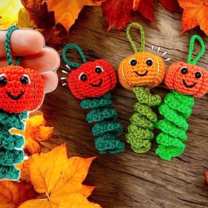 May include: Crocheted pumpkin decorations in orange, red, and green, with smiling faces and green spiral stems. The ornaments are displayed on a wooden surface with autumn leaves.