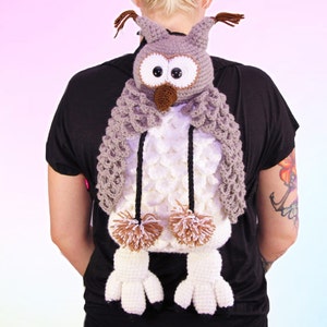 Può includere: Uno zaino a forma di gufo lavorato a uncinetto con occhi marroni, un becco marrone e pon pon bianchi e marroni. Lo zaino è indossato da una persona con una camicia nera e un tatuaggio sul braccio.
