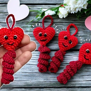 Op de afbeelding: Vier gehaakte rode hartvormige sleutelhangers met lachende gezichten. Elke sleutelhanger heeft een spiraalvormige staart.