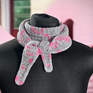 May include: A gray and pink crocheted scarf is draped over a black mannequin. The scarf features a repeating pink pattern on a gray background. The scarf is knotted at the front, showcasing its design and texture. The scarf is a cozy accessory.