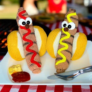 May include: Two whimsical, crocheted hot dogs with googly eyes, mustard, and ketchup on a white plate. The hot dogs have brown and white striped "meat" and are nestled in yellow buns. A red and white checkered tablecloth is in the foreground.