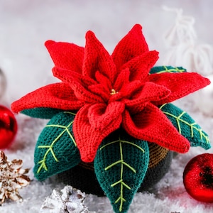 以下が含まれることがあります： 赤い花びらと緑の葉を持つ、茶色の鉢植えのクロシェのポインセチア。植物は白い模造雪と赤と銀のオーナメントに囲まれている