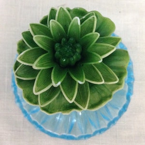 K&ouml;nnte beinhalten: Eine gr&uuml;ne Keramikblume mit einem blauen Sockel. Die Blume hat viele Bl&uuml;tenbl&auml;tter und eine Mitte, die wie eine Knospe aussieht.