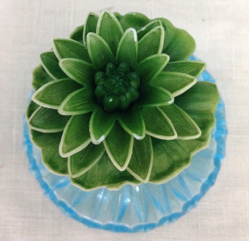 K&ouml;nnte beinhalten: Eine gr&uuml;ne Keramikblume mit einem blauen Sockel. Die Blume hat viele Bl&uuml;tenbl&auml;tter und eine Mitte, die wie eine Knospe aussieht.