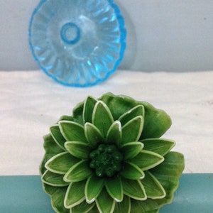 K&ouml;nnte beinhalten: Ein gr&uuml;ner, blumenf&ouml;rmiger Dekorknopf mit wei&szlig;em Rand. Der Knopf ist aus Kunststoff und hat eine strukturierte Oberfl&auml;che.