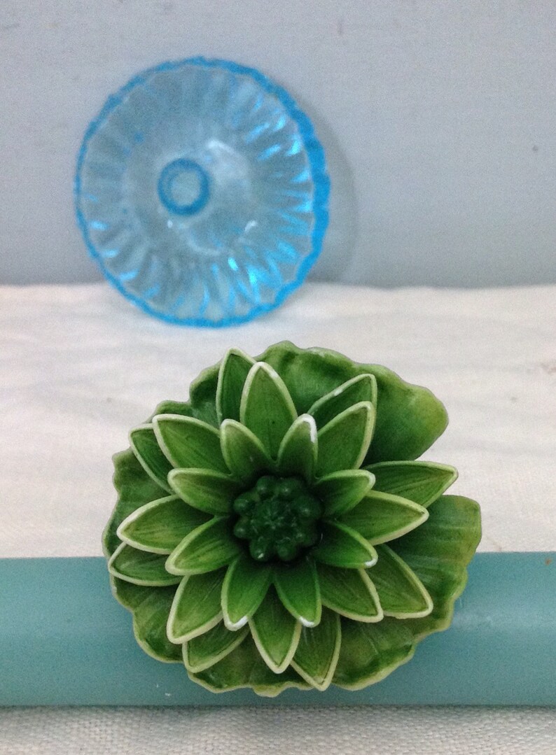K&ouml;nnte beinhalten: Ein gr&uuml;ner, blumenf&ouml;rmiger Dekorknopf mit wei&szlig;em Rand. Der Knopf ist aus Kunststoff und hat eine strukturierte Oberfl&auml;che.