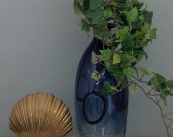 Könnte beinhalten: Eine blaue Glasvase mit strukturierter Oberfläche und einer goldenen Muschel-Skulptur auf einer weißen Oberfläche. Die Vase ist mit grünem Efeu gefüllt.