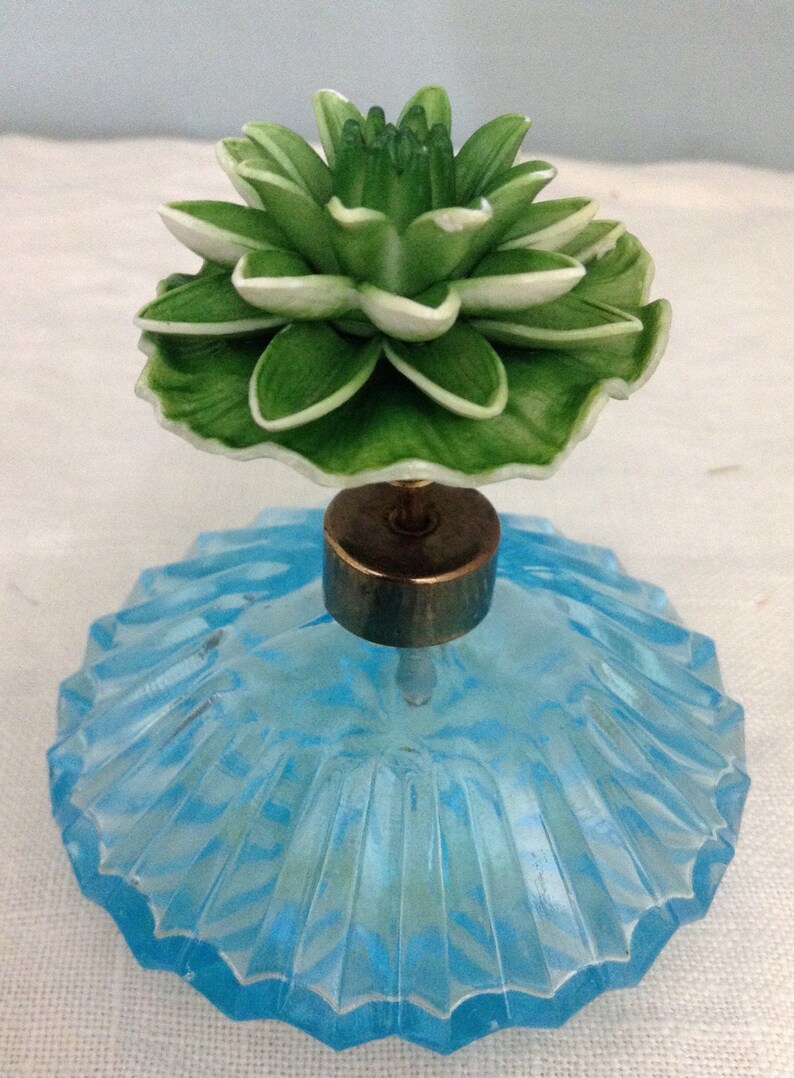 K&ouml;nnte beinhalten: Eine Vintage-Parf&uuml;mflasche aus blauem Glas mit einem gr&uuml;nen und wei&szlig;en Blumenverschluss.