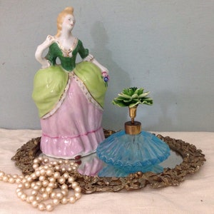 K&ouml;nnte beinhalten: Eine antike Porzellanfigur einer Frau in einem gr&uuml;n-rosa Kleid, die eine Blume h&auml;lt, steht auf einem spiegelnden Tablett mit einem blauen Glasparf&uuml;mflakon mit einem gr&uuml;nen Blumenverschluss und einer Perlenkette.