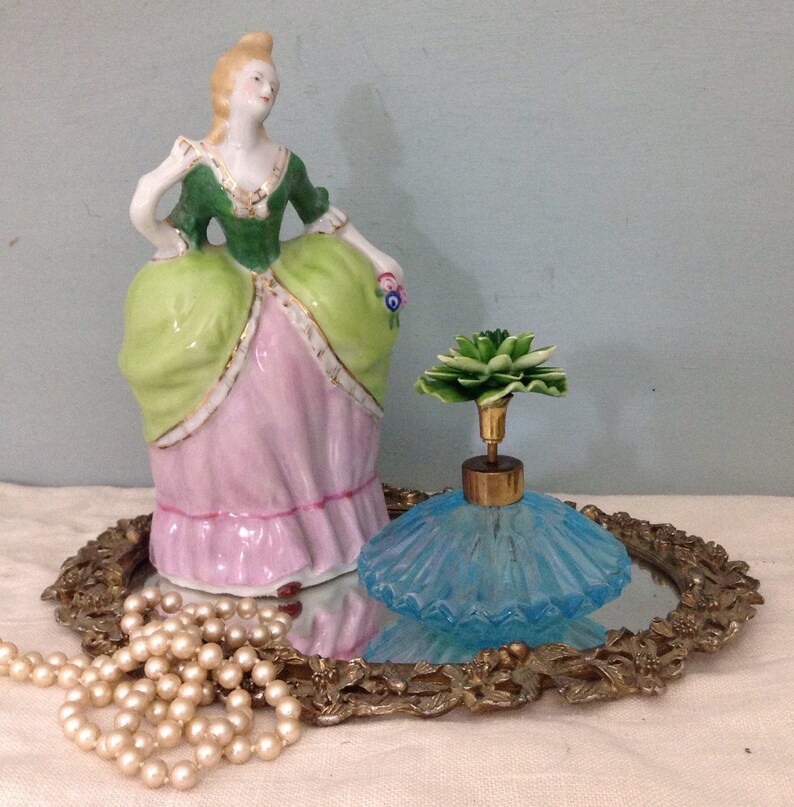 K&ouml;nnte beinhalten: Eine antike Porzellanfigur einer Frau in einem gr&uuml;n-rosa Kleid, die eine Blume h&auml;lt, steht auf einem spiegelnden Tablett mit einem blauen Glasparf&uuml;mflakon mit einem gr&uuml;nen Blumenverschluss und einer Perlenkette.