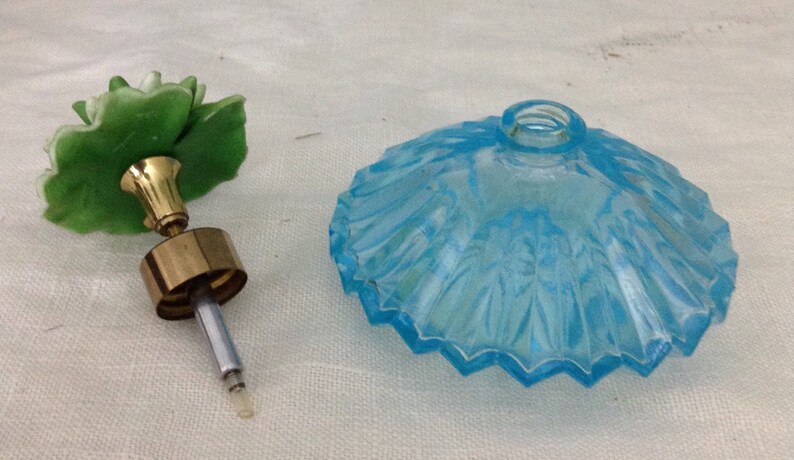 K&ouml;nnte beinhalten: Eine antike Glasparf&uuml;mflasche mit einem blauen Glasdeckel und einem gr&uuml;nen Glasblumenverschluss. Die Flasche hat ein zartes, gewelltes Design.