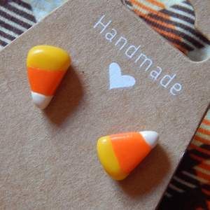 Puede incluir: Un par de pendientes artesanales de caramelos de maíz. Cada pendiente presenta un diseño amarillo, naranja y blanco, que se asemeja al clásico dulce de Halloween. Los pendientes se exhiben en una tarjeta marrón con la palabra "Handmade" y un corazón.
