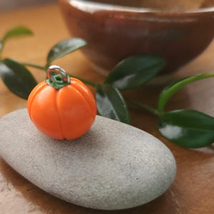 May include: A miniature orange pumpkin charm with a green stem and silver loop rests on a grey stone. The charm is round with vertical grooves. Green leaves and a brown bowl are in the background.