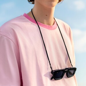 Könnte beinhalten: Ein hellrosa kurzärmeliges T-Shirt mit einem dunkelrosa Rundhalsausschnitt. Eine schwarze Sonnenbrille hängt an einer dünnen schwarzen Kordel um den Hals und liegt auf der Brust. Die Sonnenbrille hat ein rechteckiges Rahmendesign.