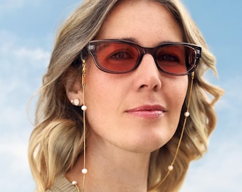 Op de afbeelding: Transparante donkergrijze zonnebril met roodachtig getinte glazen, bevestigd aan een fijne goudkleurige metalen ketting. De ketting is voorzien van kleine, gelijkmatig verdeelde witte kralen en wordt met goudkleurige karabijnsluitingen aan het montuur bevestigd.