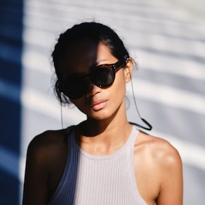 Könnte beinhalten: Eine Frau trägt ein hellgraues Tanktop und eine dunkelbraune Sonnenbrille mit einem schwarzen Band. Die Sonnenbrille bedeckt den größten Teil ihres Gesichts. Sie steht vor einer weißen Wand mit Schatten.