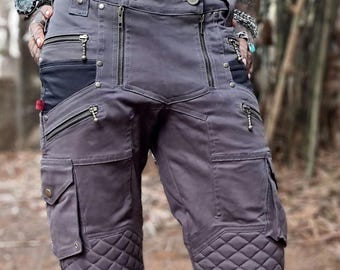 May include: Gray cargo shorts with multiple pockets, zippers, and quilted accents on the lower legs. The shorts have a utilitarian design, featuring a button closure and a black pocket. The model is wearing black boots and socks.