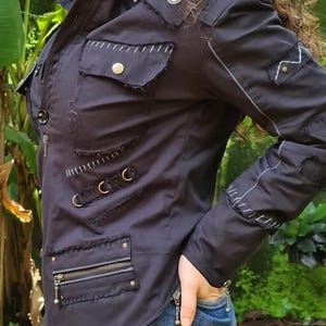 Puede incluir: Una chaqueta negra con bordes deshilachados, costuras blancas en contraste, m&uacute;ltiples bolsillos con cremalleras y botones de presi&oacute;n met&aacute;licos, y detalles de herrajes en tono plateado. Incluye mangas largas con pu&ntilde;os de cremallera y cuello alto.