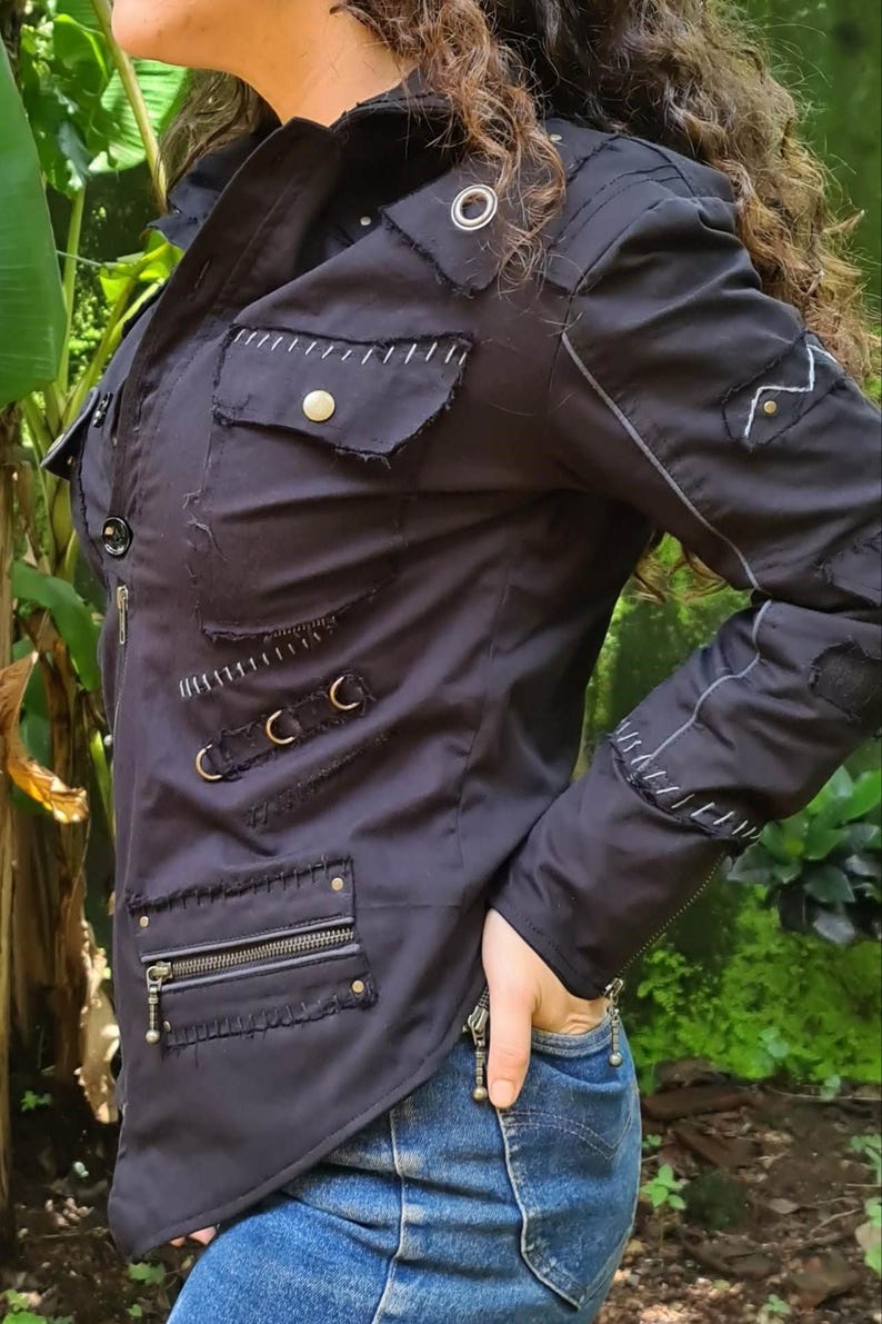 Puede incluir: Una chaqueta negra con bordes deshilachados, costuras blancas en contraste, m&uacute;ltiples bolsillos con cremalleras y botones de presi&oacute;n met&aacute;licos, y detalles de herrajes en tono plateado. Incluye mangas largas con pu&ntilde;os de cremallera y cuello alto.