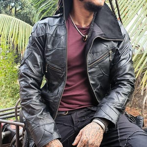 Puede incluir: Una chaqueta de cuero negra con diseño de parches de múltiples paneles cosidos, cuello alto y cremalleras de metal plateado en el frente y el pecho. La chaqueta se lleva sobre una camiseta color granate con un collar de cadena plateada.