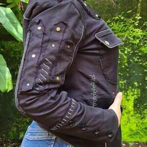 Puede incluir: Una chaqueta negra con cuello alto y costuras blancas en contraste, tachuelas met&aacute;licas doradas en las mangas, un bolsillo en el pecho con solapa y botones negros en los pu&ntilde;os. La chaqueta incluye costuras decorativas en zigzag de color blanco.