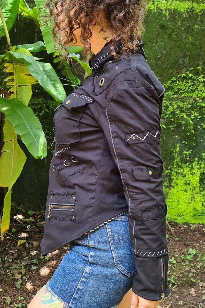 Puede incluir: Una chaqueta negra con dobladillo asim&eacute;trico, m&uacute;ltiples bolsillos con cremalleras y solapas, ojales met&aacute;licos y costuras decorativas blancas a lo largo de las costuras. La chaqueta incluye detalles de tela desgastada y una cremallera frontal.