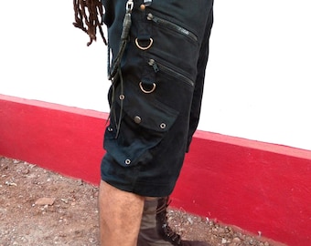 May include: Black cargo shorts with multiple pockets and zippers, paired with brown lace-up boots. The shorts feature metal hardware and a silver carabiner clip. The boots have a rugged design, suitable for outdoor activities.